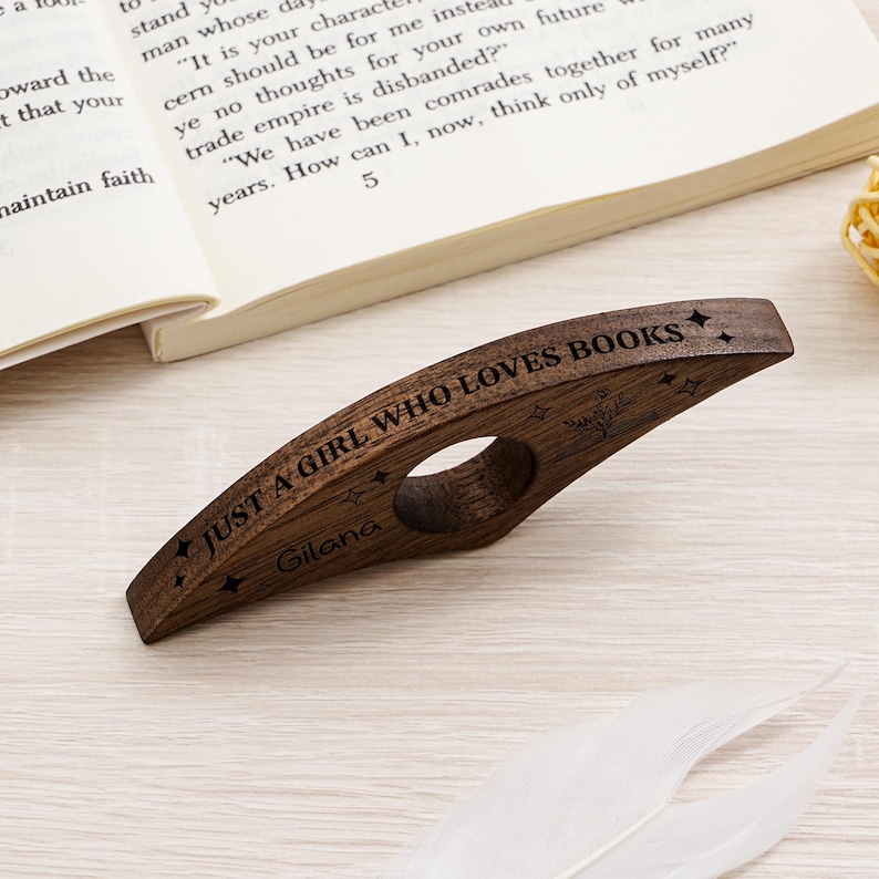 May include: A wooden book page holder with the text "JUST A GIRL WHO LOVES BOOKS" and the name "Gilana" engraved on it. The dark brown holder has a circular hole for the thumb and is placed on a light-colored surface next to an open book.