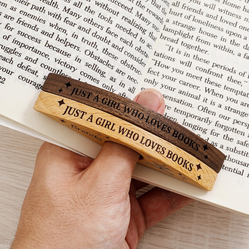 May include: Two wooden book page holders, one light and one dark, are held over a book. The text "JUST A GIRL WHO LOVES BOOKS" is engraved on each. The light wood holder has the name "Brenda" engraved.