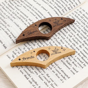 May include: Two wooden thumb page holders with personalized names, "Gilana" and "Brenda", are displayed on an open book. The dark brown and light wood-toned accessories feature a star and floral design, ideal for book lovers.