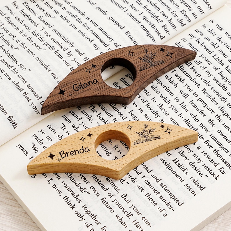 May include: Two wooden thumb page holders with personalized names, "Gilana" and "Brenda", are displayed on an open book. The dark brown and light wood-toned accessories feature a star and floral design, ideal for book lovers.