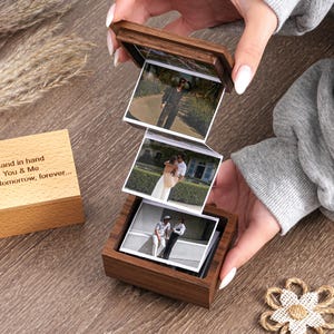 May include: A dark wood photo box with a hinged lid, displaying three stacked photographs. The box is held open, revealing the images. A smaller wooden box with text is visible to the left. A decorative flower is in the bottom right.