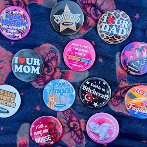 Puede incluir: Una colección de coloridas chapas con varios diseños y textos. Algunas chapas presentan frases como "I ❤️ UR DAD" y "Don't Hate Yourself, That's Homophobic." Otros diseños incluyen un gato, un diablo y una estrella.