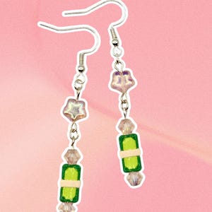 May include: A pair of dangle earrings featuring silver-toned hooks, each adorned with a translucent star-shaped bead, a clear faceted bead, and a rectangular green and cream-colored bead. The design is symmetrical, showcasing a playful arrangement of colorful beads suspended from metal findings, creating a distinct visual contrast between the star elements and the rectangular components.