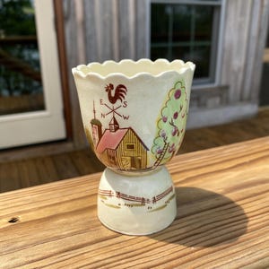 May include: Cream-colored ceramic egg cup with a scalloped rim. The cup features a farm scene with a red-roofed barn, a rooster weather vane, and a tree with pink fruit. The base has a painted fence design.