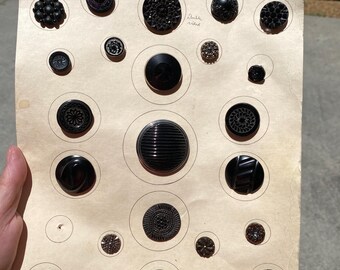 May include: A collection of various black buttons of different sizes and designs arranged on a tan paper. The buttons include floral, ribbed, and textured patterns. Some buttons have a glossy finish.