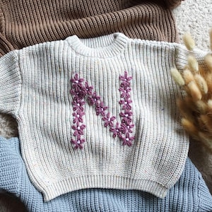 Könnte beinhalten: Ein cremefarbener Strickpullover mit einem großen, lila bestickten Buchstaben "N" aus Blumen. Der Pullover ist aus weichem, strukturiertem Garn gefertigt.