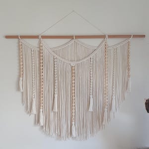 May include: A cream-coloured macrame wall hanging suspended from a horizontal wooden dowel. The design features intricate knotted patterns, vertical strands of light-coloured wooden beads, and multiple hanging tassels along the bottom edge.