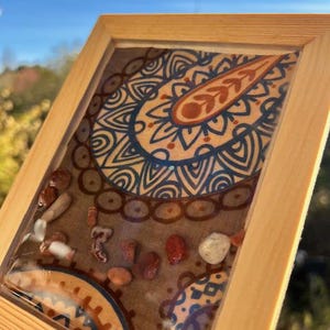 Puede incluir: Obra de arte enmarcada en madera con un diseño de cachemira en tonos azules, marrones y naranjas. Pequeñas piedras están incrustadas en resina. El marco es de madera clara. La obra de arte es sostenida por una mano.