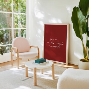Può includere: Un'opera d'arte da parete rettangolare incorniciata con uno sfondo rosso intenso e il testo scritto a mano in bianco "joy is in the simple moments" al centro, posizionata accanto a una sedia imbottita rosa chiaro e un tavolino rotondo bianco.