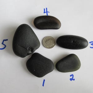 May include: Five dark, smooth beach stones of varying shapes and sizes are arranged around a dime on a white surface. The stones are primarily black, with some showing hints of green or brown. The dime provides a size reference.