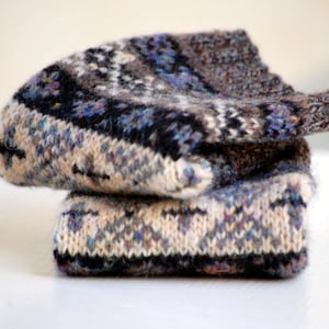 May include: A stack of two knitted hats. The hats feature a pattern of cream, black, blue, and brown. The hats are made of a soft, textured yarn. The hats are folded and stacked on top of each other.