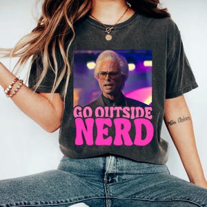 Pode incluir: Uma camiseta preta com um visual desbotado apresenta um gráfico de um homem com cabelo branco e óculos. O gráfico é cercado por um retângulo rosa com o texto "GO OUTSIDE NERD" em rosa.
