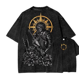 Puede incluir: Dos camisetas gráficas negras. La camiseta más grande presenta una ilustración detallada de un caballero con una espada, rodeado de flores y una calavera, sobre un fondo de rayos de sol dorados. La camiseta más pequeña muestra un emblema dorado. Ambas camisetas tienen un lavado vintage.