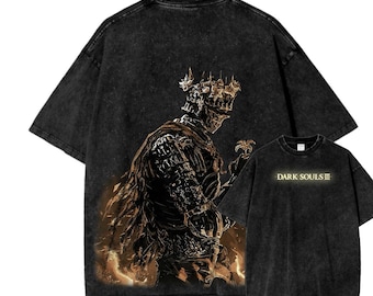 May include: Dark gray t-shirt featuring a detailed illustration of a knight with a crown and armor. The design is in shades of gold and brown. The text "DARK SOULS III" is printed on a smaller shirt in the image.