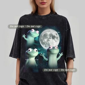 Puede incluir: Camiseta gris oscuro con un diseño caprichoso de tres ranas de dibujos animados y una luna llena de fondo. Las ranas son de color verde claro con ojos grandes. El texto "Do not copy / Do not copy" está impreso en la camiseta.