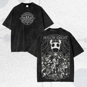 May include: Black t-shirt with white graphics on the front and back. The front displays a circular design with a central character. The back features the text "HOLLOW KNIGHT" above a detailed illustration of various characters.