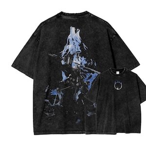 Puede incluir: Camisetas negras con lavado ácido con un diseño gráfico. La camiseta más grande presenta una ilustración azul y blanca de un lobo y un guerrero. La camiseta más pequeña tiene un diseño circular blanco.