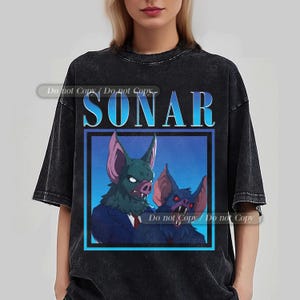 May include: Dark gray t-shirt featuring a graphic of two bat-like creatures with the word "SONAR" in blue above them. The creatures have green and purple accents, set against a blue background.