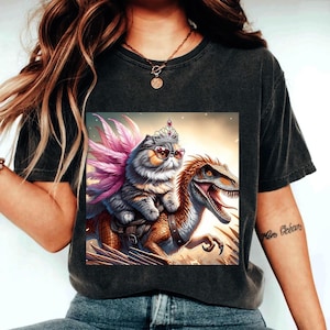 Puede incluir: Una camiseta negra con un estampado gráfico de un gato con una corona y gafas de sol montando un dinosaurio. El gato tiene plumas rosas en la espalda.