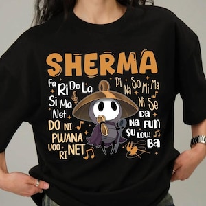May include: Black t-shirt featuring the word "SHERMA" in orange, with a cartoon character wearing a hat and holding a musical instrument. Musical notes and other text surround the character. The shirt is a casual wear item.