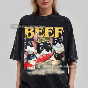 Puede incluir: Camiseta negra con un diseño de dibujos animados de cinco perros y la palabra "BEEF" en amarillo. Los perros tienen pelaje blanco y negro, uno de ellos con capa y máscara. El fondo incluye rayos.