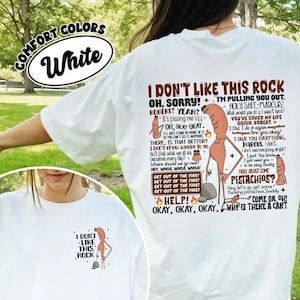 Puede incluir: Camiseta blanca con un gráfico humorístico. El diseño incluye la frase "I don't like this rock" y personajes de dibujos animados, junto con otros elementos de texto y frases.