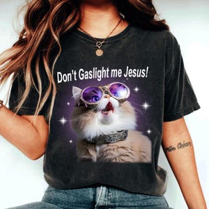 Puede incluir: Una camiseta negra con un gato blanco que lleva gafas de sol y un collar de joyas. El texto "Don't Gaslight me Jesus!" está impreso en la camiseta.