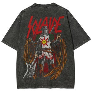 May include: Dark gray t-shirt featuring a graphic of a knight with a sun emblem, wings, and an axe. The word "SOLAIRE" is printed in red across the top. The shirt has a vintage, distressed look.