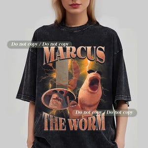 Puede incluir: Camiseta negra de gran tamaño con un gráfico de un personaje de gusano y el texto "MARCUS THE WORM". El diseño incluye rayos y un estilo retro. La camiseta tiene un aspecto vintage y descolorido.
