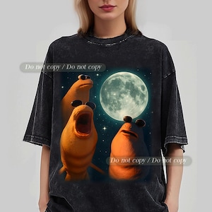 May include: Dark grey t-shirt with a quirky design of three orange creatures wearing sunglasses, set against a starry night sky and a full moon. The text "Do not copy / Do not copy" is printed on the shirt.