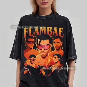 May include: Black t-shirt featuring a graphic of a man wearing pink sunglasses, surrounded by other figures. The word "FLAMBAE" is printed in orange and yellow above the central image. The shirt has a vintage, distressed look.