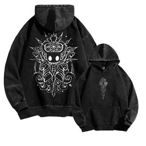 Puede incluir: Dos sudaderas con capucha negras. La sudadera más grande presenta un gráfico blanco en la espalda de un personaje con corona y detalles ornamentados. La sudadera más pequeña tiene un gráfico blanco a juego en la parte delantera. Ambas sudaderas tienen un aspecto vintage.