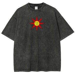 May include: Dark gray t-shirt featuring a stylized sun graphic with a yellow face and red rays. The shirt has a vintage, washed appearance and a classic crew neck design. A casual, unisex tee.