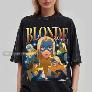 Puede incluir: Camiseta negra con un diseño retro. Presenta una mujer rubia con una máscara azul y un top amarillo, con las palabras "BLONDE Blazer" en oro. El diseño tiene un aspecto vintage y desgastado.
