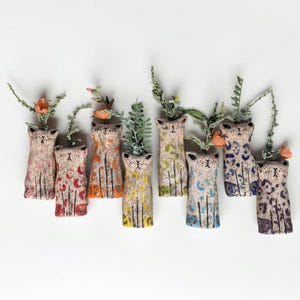 May include: A collection of seven ceramic cat-shaped bud vases, each featuring a unique speckled finish and distinct colorful patterns. Each cat figure has a small opening at the top containing a sprig of dried flowers or greenery.