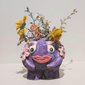 May include: A purple ceramic creature planter with a glossy finish, featuring large white eyes, pink lips, and pink flower accents on the sides. The figure has two small arms and legs, and it is filled with a bouquet of yellow and orange wildflowers.
