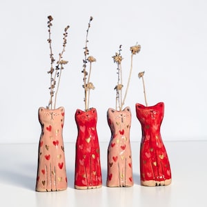 Könnte beinhalten: Vier keramische Vasen in Katzenform in Rosa- und Rottönen, verziert mit goldenen Herzakzenten. Jede Vase enthält getrocknete Blumenstiele. Die Vasen sind auf einer weißen Oberfläche angeordnet und bilden eine charmante und dekorative Präsentation.