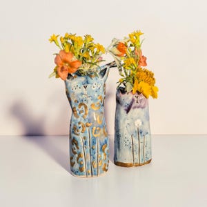 Könnte beinhalten: Zwei hellblaue, katzenförmige Keramikvasen mit bemalten Details. Die größere Vase hat goldene Leopardenflecken, während die kleinere violette Akzente und ein weißes Blumenmuster aufweist. Beide Vasen sind mit gelben und orangen Blumen gefüllt.