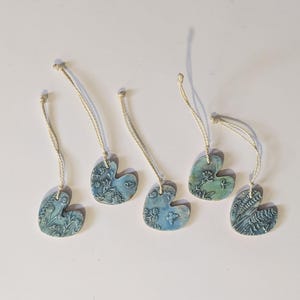 May include: Five blue ceramic heart-shaped ornaments with floral designs and cream-colored string hangers. The ornaments have a textured surface and varying shades of blue and green. The ornaments are arranged on a white surface.