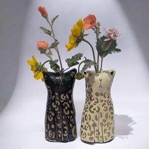 May include: Two ceramic cat-shaped vases, one black with gold spots and the other cream with brown spots, each holding colorful flowers. The vases have a whimsical design, with the cat's face and paws detailed. The flowers include yellow, orange, and pink blooms.
