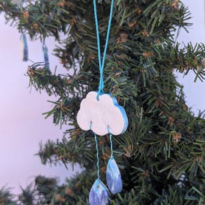 Może przedstawiać: Ręcznie robiona ozdoba choinkowa w kształcie chmury z niebieskimi akcentami i dwoma wiszącymi zawieszkami w kształcie kropli deszczu. Ozdoba jest biała z niebieskimi obwódkami, zawieszona na jasnoniebieskim sznurku.