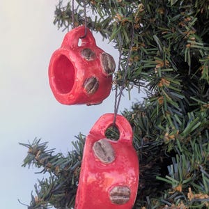 May include: Two red ceramic coffee cup ornaments with brown coffee bean accents hang from a Christmas tree. The ornaments are glossy and have a hole at the top for hanging. The coffee cups are a festive holiday decoration.