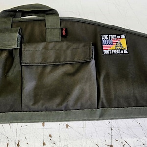 Könnte beinhalten: Eine olivgrüne Gewehrtasche mit mehreren Außentaschen und einem Tragegriff. Die Tasche hat einen Aufnäher mit der amerikanischen Flagge und dem Text "LIVE FREE OR DIE, DON'T TREAD ON ME". Ein kreisförmiger Aufnäher mit einem spartanischen Helm ist ebenfalls sichtbar.