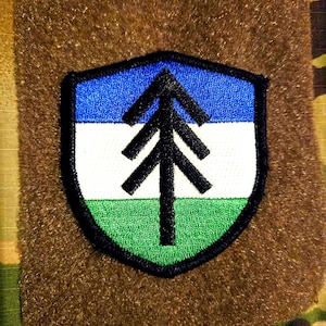 May include: Embroidered patch with a shield design and a black border. The shield is split into three horizontal sections: blue, white, and green. A black tree graphic is in the centre, set against a brown background.