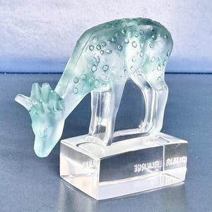 May include: A frosted light green glass figurine of a grazing deer with a spotted pattern on its back. The deer stands on a clear, rectangular glass base that features etched text on the front side, displaying a smooth and translucent finish.