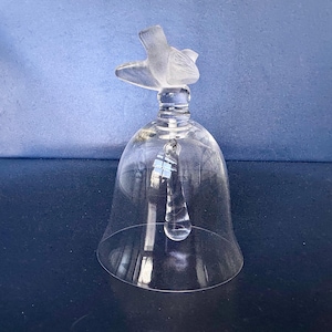 May include: A clear glass bell with a bird-shaped handle. The bell has a teardrop-shaped clapper and is set against a blue background. The bird is frosted, and the bell is transparent.