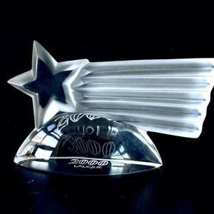 May include: A clear glass sculpture of a shooting star with a five-pointed star at the front. The star has a frosted finish and the tail of the shooting star is made of clear glass with a frosted finish. The base of the sculpture is a clear glass oval with the words "2000 Lalique" engraved on it.