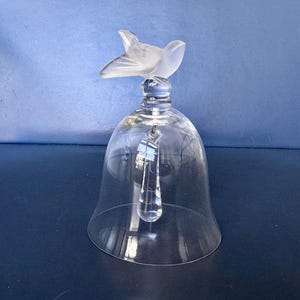 May include: A clear glass bell with a frosted bird figurine on top. A clear, teardrop-shaped crystal hangs inside the bell. The bell is sitting on a dark surface.