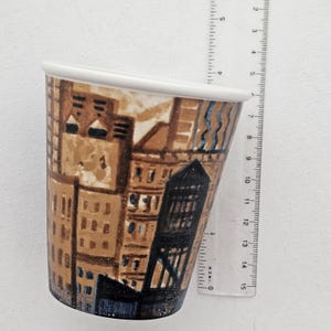 Puede incluir: Una taza de cer&aacute;mica con un dise&ntilde;o de paisaje urbano. La taza tiene un borde blanco y una forma c&oacute;nica. La obra de arte representa edificios marrones y azules. La taza mide aproximadamente 13 cm de altura.
