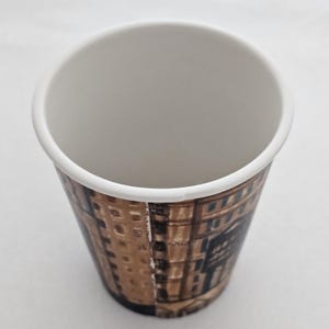 Puede incluir: Una taza de caf&eacute; de papel desechable con interior y borde blancos. El exterior presenta un dise&ntilde;o arquitect&oacute;nico marr&oacute;n y beige. La taza est&aacute; vac&iacute;a y se encuentra sobre una superficie blanca.
